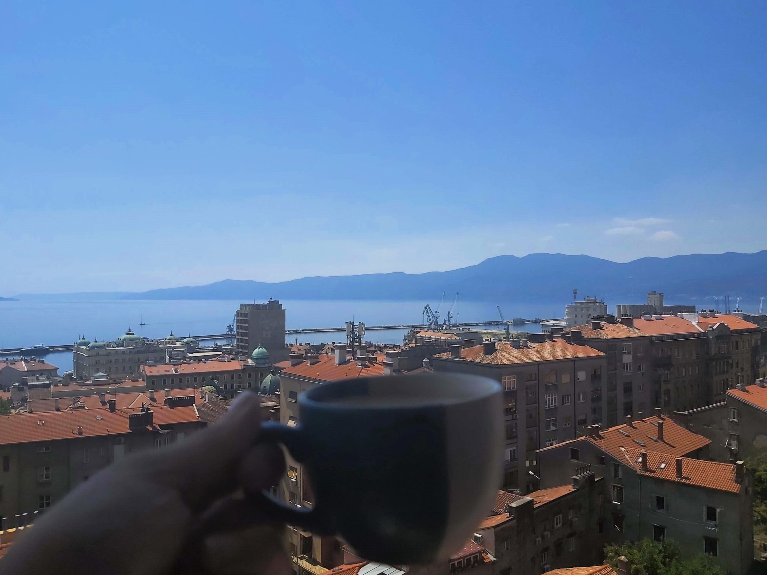 Wandering Lewis holds an espresso up against the mountainous sea view from her apartment view in Rijeka, Croatia