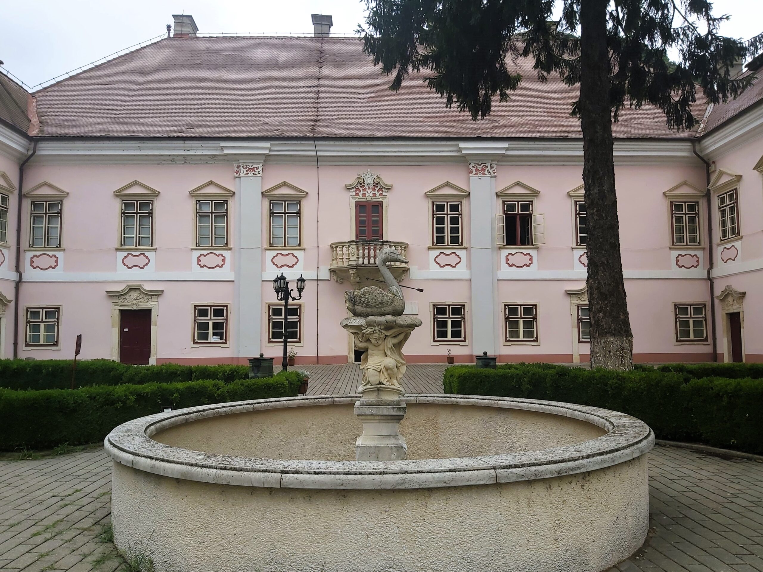 A carved fountain of a goose pierced with an arrow and pink Museum of Dacian and Roman Civilization building in the background in Deva, Romania