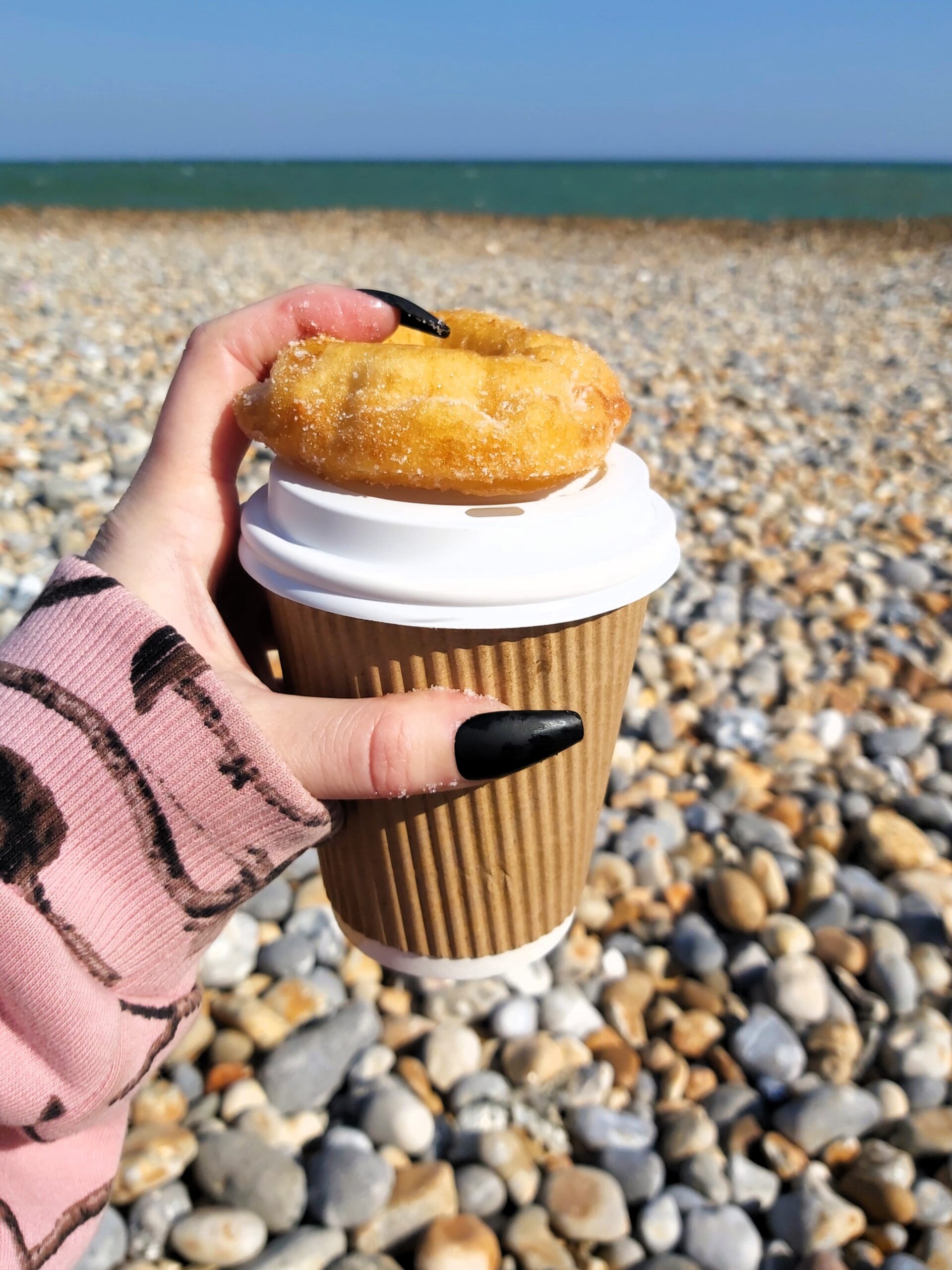 Wandering Lewis, with long black nails, holds a donut and coffee on the beach at Hastings, England.