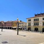 A colourful shot of the buildings in Piazza Ferretto (or Piazza Maggiore). Buildings of different colours, including yellow, peach, brown and green, are set off against a blue sky. Location is Mestre, Italy.