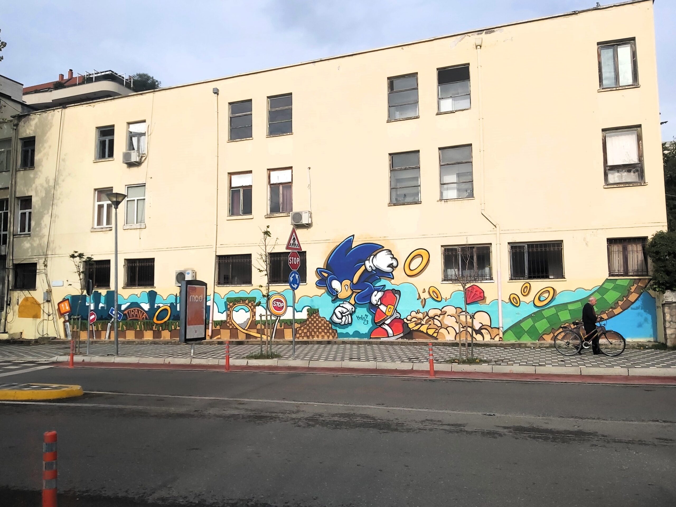 Sonic the Hedgehog mural on a building in Tirana, Albania, with a man and his bike passing.