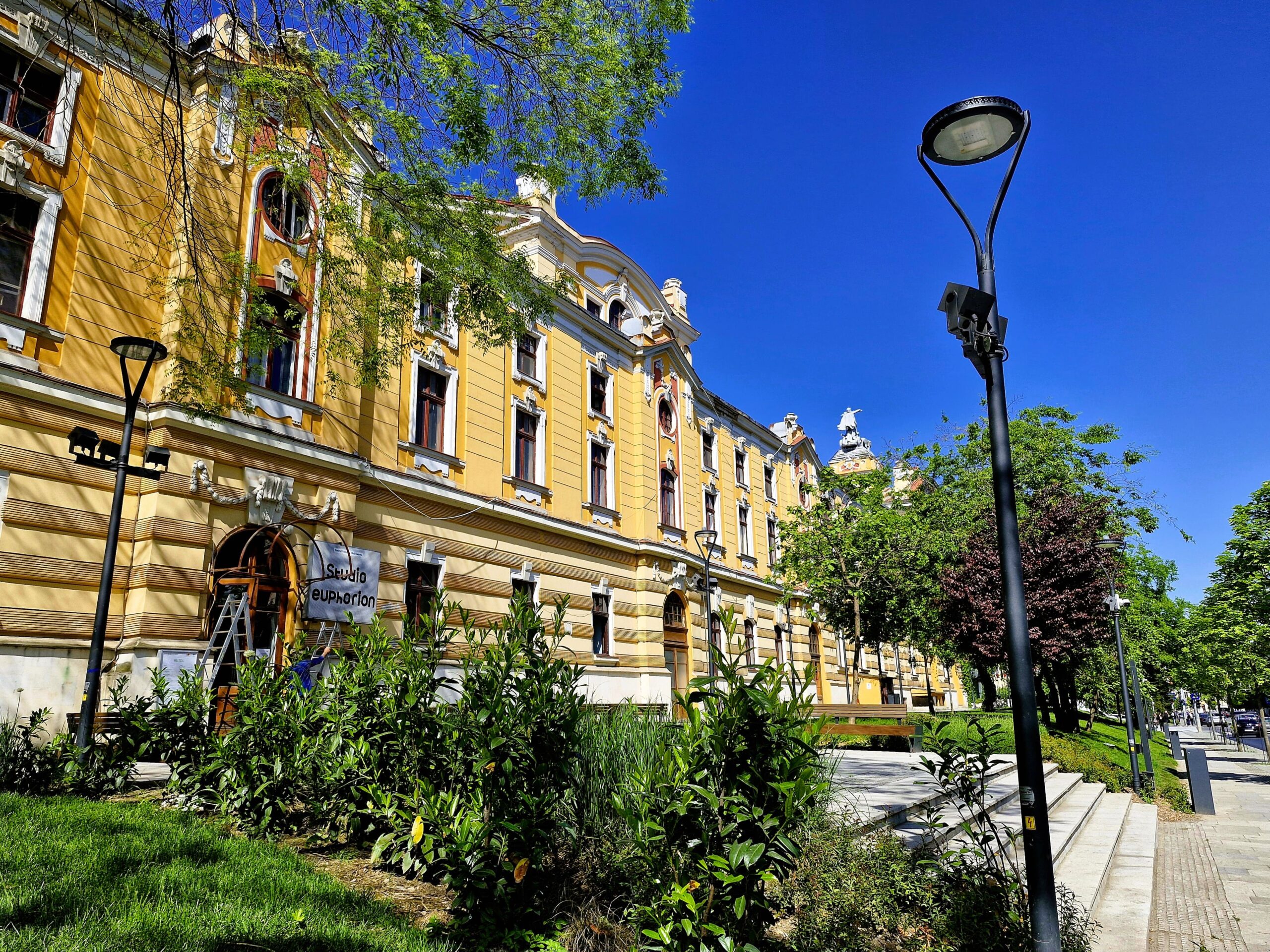 The yellow Opera building in Cluj-Napoca with contrasting green trees and clear blue sky.