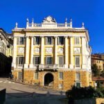 A very yellow building with pretty, ornate plaques and statues sits proud in a street of Bergamo.