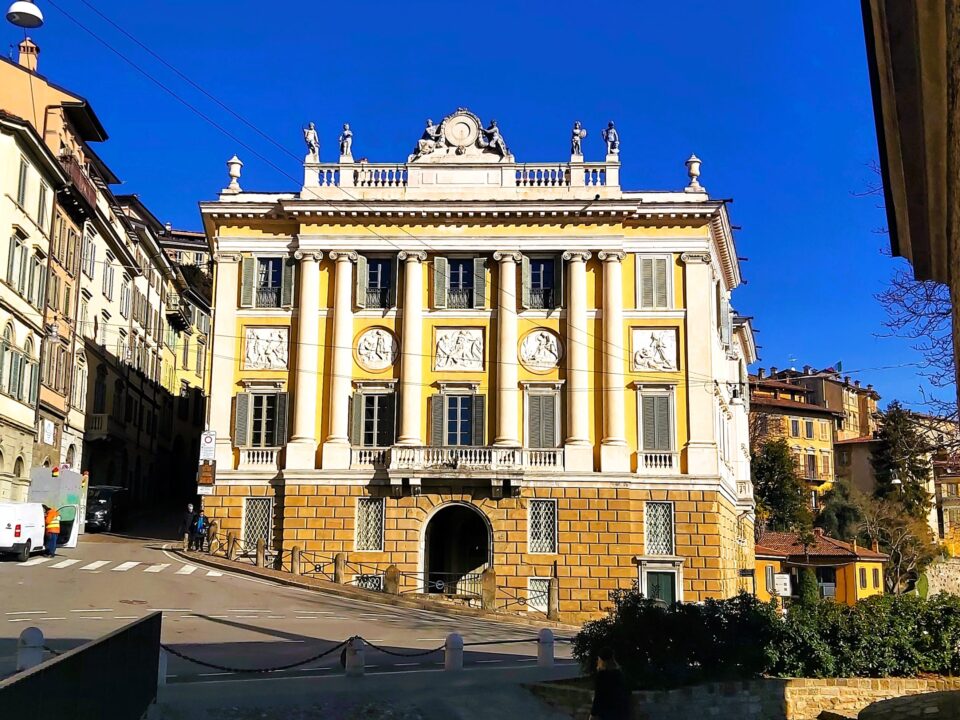 A very yellow building with pretty, ornate plaques and statues sits proud in a street of Bergamo.