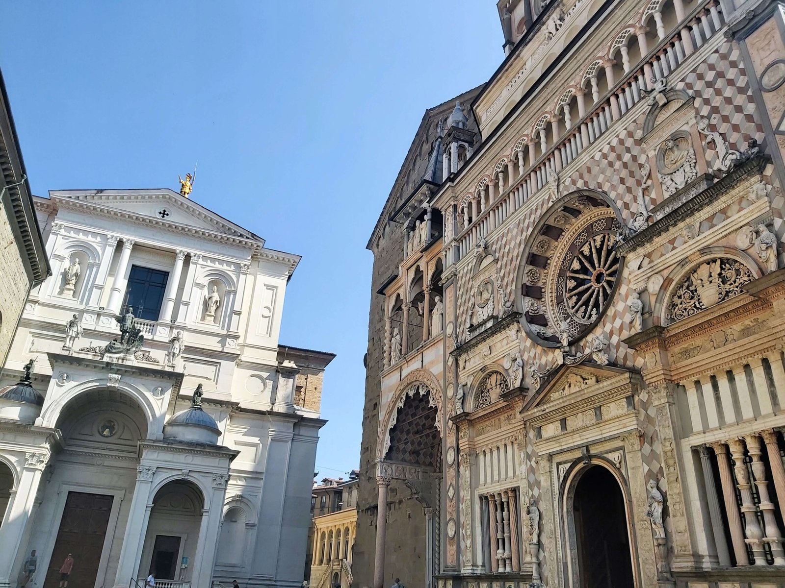 A beautiful pair of old buildings, a bright white church, next to an ornate fascia in many shades of brown and cream. Bergamo, Italy